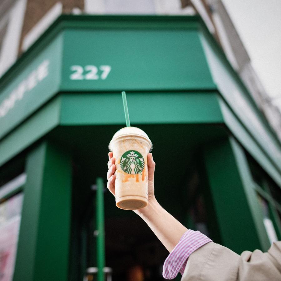 Hand holding a Starbucks takeaway cup in front of a green Starbucks building