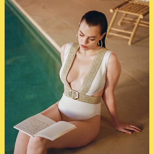 Model in white and gold swimsuit with slicked back hair reads at the poolside